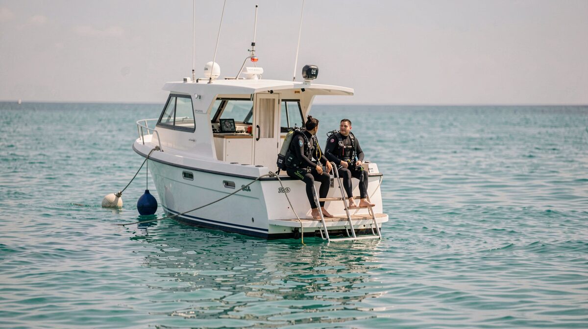 A small white dive boat tied to a mooring buoy on flat turquoise water off a lava coastline, stern ladder down, mid-morning light.